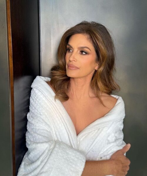 Modelka Cindy Crawford w pełnym makijażu, eleganckiej fryzurze, okrywa się białym szlafrokiem. Patrzy w bok i stoi na tle szarej ściany.