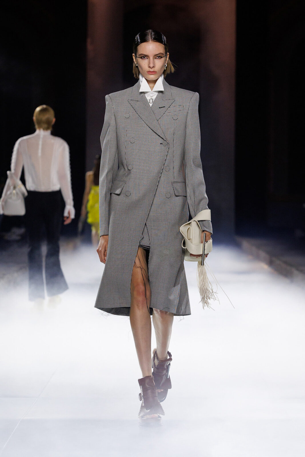 Modelka prezentuje stylizację złożoną z eleganckiego płaszcza w drobną kratkę o kroju oversize, zapinanego dwurzędowo, z delikatnym rozcięciem z przodu. Pod płaszczem widoczny jest element prześwitującej tkaniny. Stylizację dopełniają brązowe botki z charakterystycznymi wycięciami oraz torebka w jasnym odcieniu z frędzlami, trzymana w dłoni. Tło stanowi efektowne, mglisto-podświetlone wnętrze wybiegowe, które nadaje całości dramatycznego charakteru.