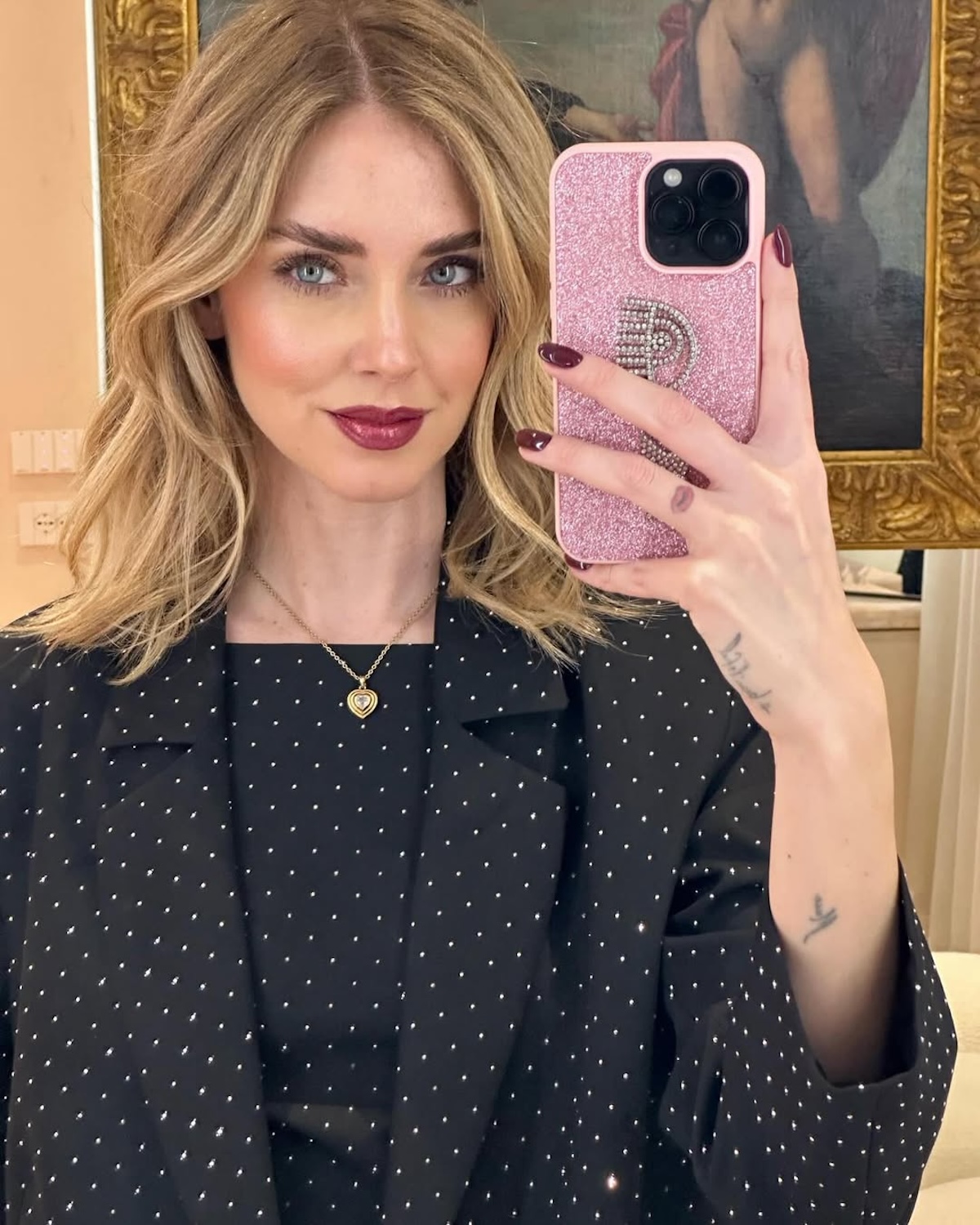 Chiara Ferragni torebki: W czarnym eleganckim garniturze, prezentująca różowe etui na telefon z błyszczącym logo oka. Stylizacja łączy nowoczesność i subtelne detale, a selfie idealnie oddaje jej rozpoznawalny styl.