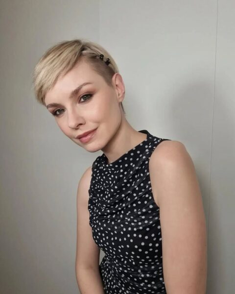 Modelka o blond włosach ściętych w stylu pixie cut z grzywką na bok. Z jednej strony ma włosy podpięte spineczkami. Ubrana jest w czarny top w białe groszki.