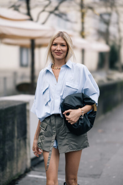 Stylizacja casual-chic – oversize’owa koszula w chłodnym błękicie z szerokimi kieszeniami zestawiona z krótką spódnicą w oliwkowym odcieniu oraz dużą, czarną torbą typu pouch. Delikatna biżuteria w złocie dodaje elegancji.