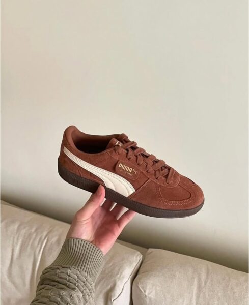 Brązowe sneakersy Puma Palermo w kolorze ciemnego miodowego brązu z białym logo i paskiem, wykonane z zamszu. Trzymane w ręku nad sofą z jasną tapicerką, na tle minimalistycznej, jasnobeżowej ściany.