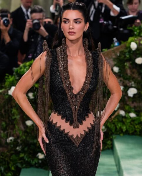 Met Gala 2025:Modelka ubrana w elegancką, zdobioną suknię z głębokim dekoltem i misternymi haftami w odcieniach brązu. Włosy zaczesane do tyłu, a jej wyraz twarzy pełen pewności siebie. W tle tłum i dekoracje przypominające ogród.