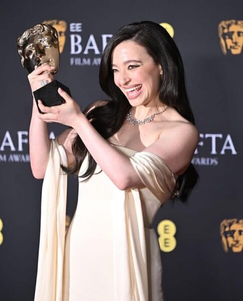 Laureatka BAFTA 2025 z radością prezentuje swoją nagrodę. Ubrana w elegancką kremową suknię z odkrytymi ramionami, z diamentowym naszyjnikiem na szyi, trzyma statuetkę BAFTA i uśmiecha się szeroko, emanując szczęściem. Stylizacja BAFTA 2025 podkreśla klasyczną elegancję z nutą nowoczesności.