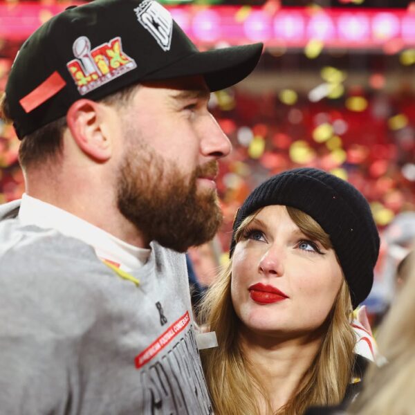 Taylor Swift w czarnej czapce z dzianiny patrzy z podziwem na mężczyznę w szarej koszulce i czapce z daszkiem, ozdobionej logo Super Bowl. W tle widać rozmyte kolorowe światła i konfetti unoszące się w powietrzu, co sugeruje świąteczną atmosferę.