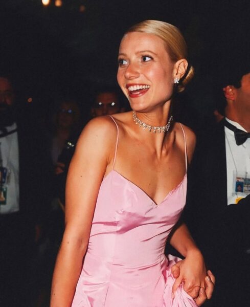 Gwyneth Paltrow uśmiechnięta podczas gali Oscarów 1999. Aktorka ma na sobie ikoniczną różową suknię z cienkimi ramiączkami od Ralpha Laurena. Stylizację uzupełniają delikatny diamentowy naszyjnik i kolczyki. Jej włosy są gładko zaczesane w elegancki kok, a makijaż subtelnie podkreśla naturalne piękno. W tle elegancko ubrani goście.