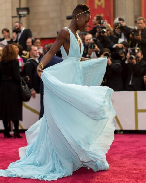 Lupita Nyong'o na czerwonym dywanie podczas Oscarów 2014. Aktorka ma na sobie zachwycającą, błękitną suknię od Prady z głębokim dekoltem i lekką, plisowaną spódnicą, która efektownie unosi się w ruchu. Stylizację dopełnia złota opaska na włosach, subtelna biżuteria i elegancka bransoletka. W tle tłum fotografów i gości gali, uwieczniających ten ikoniczny moment