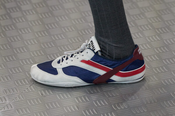 Zbliżenie na sportowy but w stylu retro z kolekcji Miu Miu na wiosnę 2025. Sneakers ma biało-granatową cholewkę z czerwonymi detalami oraz elastyczny pasek oplatający piętę. Logo marki widoczne na języku. Stylizacja uzupełniona o ciemnoszare skarpety, które dodają eleganckiego kontrastu.