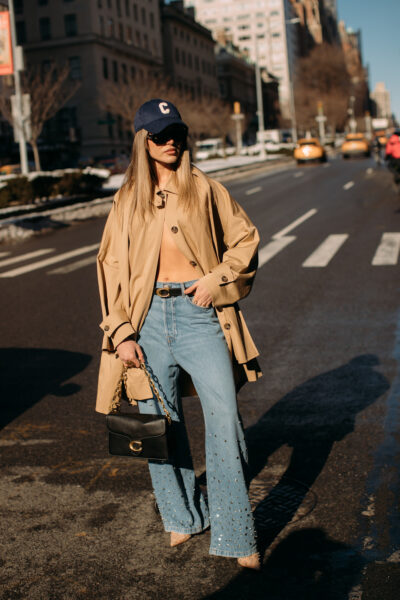 Nowojorski street style – modelka w oversizowym, beżowym trenczu noszonym luźno na ramionach, jeansach z rozszerzanymi nogawkami ozdobionymi kryształkami oraz eleganckich szpilkach. Stylizację uzupełnia czarna torebka na złotym łańcuchu oraz czapka z daszkiem.