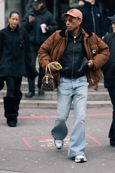 Stylizacja męska inspirowana latami 90. na ulicach Paryża. Mężczyzna w vintage brązowej kurtce, czarnym golfie z tłoczonej skóry i rozszerzanych jeansach, dopełniając look sneakersami w stylu retro, czapką z daszkiem i klasyczną torebką Louis Vuitton Speedy.