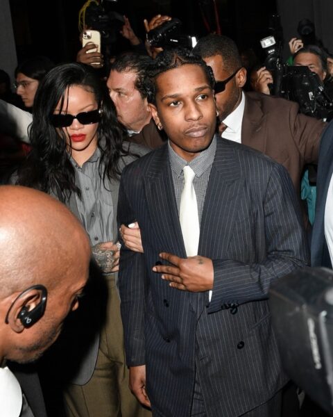 ASAP Rocky uniewinniony – raper i Rihanna wychodzą z sądu w otoczeniu ochrony i fotoreporterów. ASAP Rocky, ubrany w elegancki prążkowany garnitur, wygląda spokojnie, podczas gdy Rihanna, w szarym garniturze i ciemnych okularach, trzyma go pod ramię. Tłum dokumentuje moment kamerami i aparatami.