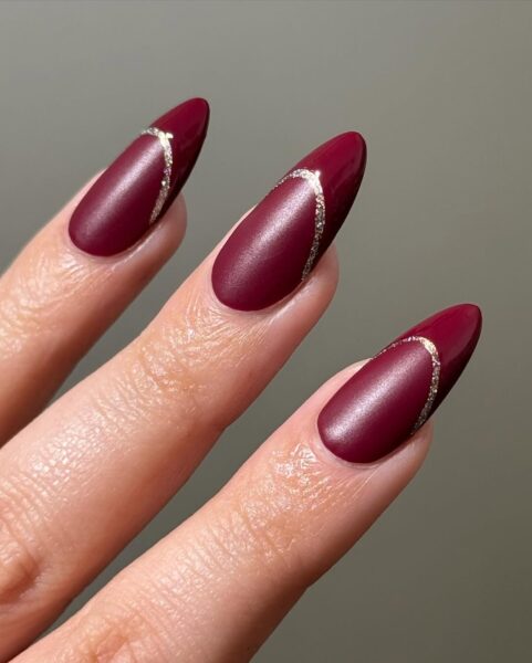 Manicure w odcieniach głębokiego wina z subtelnym złotym zdobieniem, które podkreśla wyjątkowy kształt paznokci. To idealna propozycja na paznokcie na wiosnę 2025, dodająca dłoniom elegancji i luksusowego wykończenia.