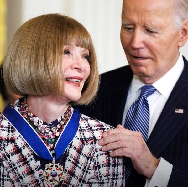 Zbliżenie na uśmiechniętą Wintour odbierającą Medal Wolności z rąk prezydenta Joe Bidena. Na szyi ma elegancki, bogato zdobiony naszyjnik, a na ramionach spoczywa niebieska wstęga z odznaczeniem. Prezydent patrzy na nią z uznaniem, lekko dotykając jej ramienia.