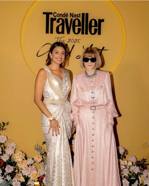 Stylowe wydarzenie Condé Nast Traveller 2025 Gold List. Dwie elegancko ubrane kobiety pozują na tle złotej ściany z logo wydarzenia. Kobieta po lewej nosi błyszczącą białą suknię, a druga – różową, długą kreację z okularami przeciwsłonecznymi i dużą biżuterią.