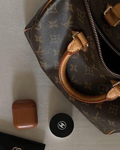 Na zdjęciu widoczna jest kultowa Louis Vuitton Speedy Bag w klasycznym wzorze monogramu na brązowym płótnie, z uchwytami i detalami wykonanymi z naturalnej skóry Vachetta, która z biegiem czasu nabiera charakterystycznej patyny. Torebka leży na eleganckiej, neutralnej powierzchni obok kilku luksusowych akcesoriów: brązowego etui na słuchawki oraz kompaktu Chanel, co podkreśla wyrafinowany styl właścicielki. Speedy Bag, będąca jednym z najbardziej rozpoznawalnych modeli Louis Vuitton, od lat pozostaje symbolem klasycznej elegancji i doskonałą inwestycją w świat vintage.