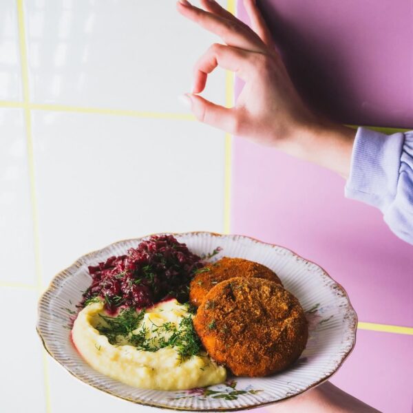 Talerz z wegańskim daniem w stylu domowej kuchni – chrupiące kotlety, kremowe puree ziemniaczane z koperkiem i duszona czerwona kapusta. W tle pastelowa ściana w odcieniach fioletu i bieli, a obok ręka pokazująca gest „OK”. Zdjęcie nawiązuje do oferty lokali z kategorii najlepsze wegańskie restauracje w Warszawie 2025.