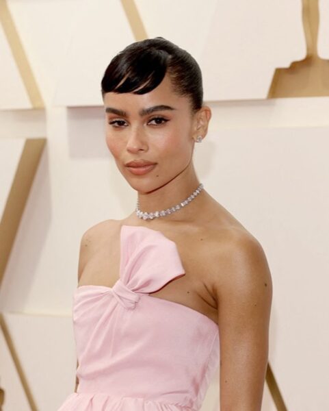 Zbliżenie na Zoë Kravitz podczas Oscarów 2022. Aktorka ma gładko zaczesane włosy z krótką grzywką i nosi delikatny diamentowy naszyjnik. Jej różowa suknia od Yves Saint Laurent ozdobiona jest asymetryczną kokardą. Stylizację dopełnia subtelny makijaż podkreślający oczy i usta