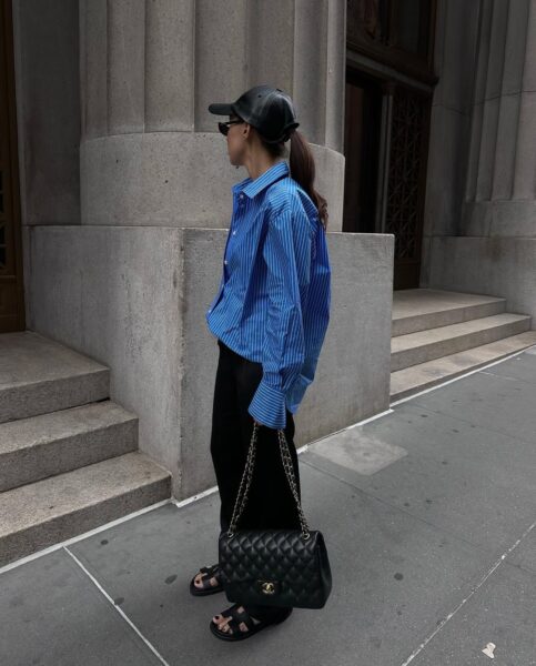 Na zdjęciu widoczna jest stylowa kobieta w minimalistycznej, eleganckiej stylizacji, która idealnie wpisuje się w estetykę współczesnego street style'u. Ma na sobie oversize’ową koszulę w biało-niebieskie paski, czarne szerokie spodnie satynowe, klasyczne czarne sandały Hermès Chypre, a całość dopełniają akcesoria: czarna skórzana czapka, okulary przeciwsłoneczne oraz ikoniczna Chanel Classic Flap Bag w czarnym kolorze z pikowaniem i złotym zapięciem CC. Stylizacja jest nowoczesna, a jednocześnie ponadczasowa – połączenie czerni i błękitu nadaje jej świeżości i miejskiego szyku. Tło, z monumentalną architekturą i betonowymi schodami, dodatkowo podkreśla klimat nowojorskiej elegancji.