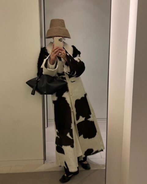 Lustrowe selfie osoby w długim, białym płaszczu w czarne łatki, co wpisuje się w trend cow print na wiosnę 2025. Stylizację dopełnia beżowy kapelusz wiadro oraz czarna torebka o geometrycznym kształcie.