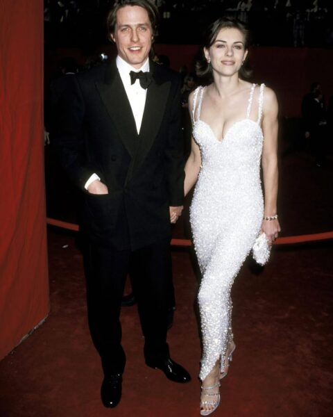 Elizabeth Hurley i Hugh Grant na czerwonym dywanie podczas Oscarów 1995. Hurley ma na sobie dopasowaną, białą suknię Versace zdobioną perłowymi aplikacjami i cienkimi ramiączkami. Stylizację uzupełniają srebrne sandałki na obcasie i mała perłowa kopertówka. Hugh Grant ubrany jest w klasyczny czarny smoking z muszką. Para trzyma się za ręce i uśmiecha do kamer.