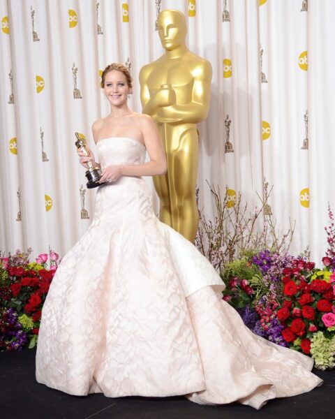 Jennifer Lawrence na ceremonii wręczenia Oscarów w 2013 roku. Aktorka pozuje ze statuetką Oscara za rolę w filmie Poradnik pozytywnego myślenia. Ma na sobie zachwycającą, jasnoróżową suknię haute couture z rozkloszowanym dołem i trenem. Stylizację uzupełniają delikatna biżuteria i upięte włosy. W tle duża złota statuetka Oscara oraz biała ścianka z logotypami ABC i dekoracją z kwiatów w odcieniach czerwieni, fioletu i zieleni.