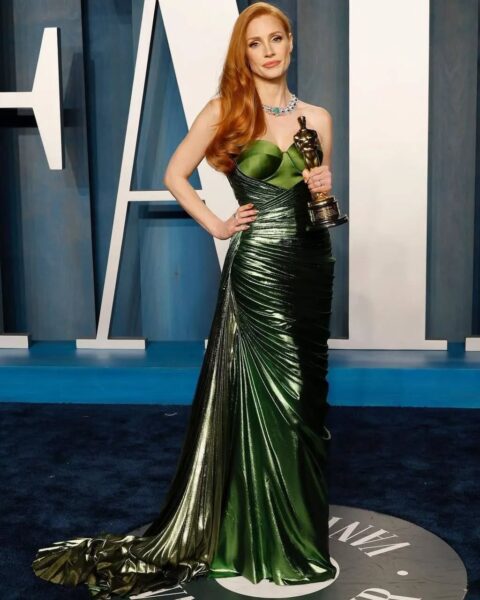 Jessica Chastain na przyjęciu Vanity Fair po Oscarach 2022. Trzyma w ręku statuetkę Oscara i pozuje w zachwycającej, metalicznie zielonej sukni o asymetrycznym kroju z drapowaniami i długim trenem. Kreacja podkreśla jej sylwetkę i współgra z jej rudymi falującymi włosami. Stylizację uzupełnia elegancka biżuteria z zielonymi kamieniami i subtelny makijaż. W tle niebieska ścianka z dużymi literami tworzącymi napis 'Vanity Fair'.