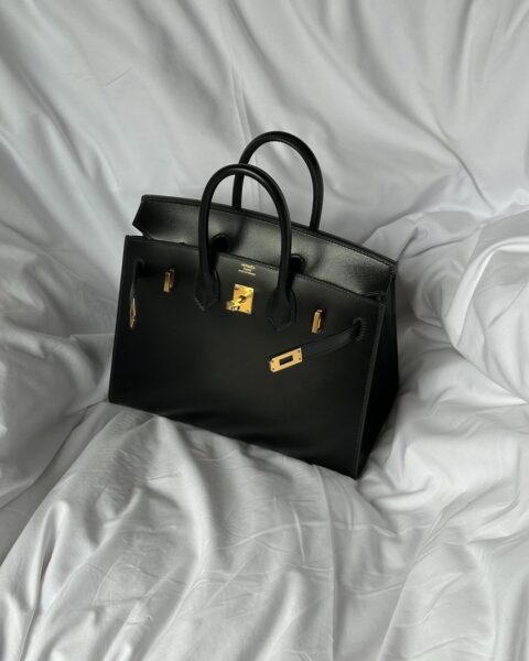 Na zdjęciu widoczna jest kultowa Hermès Birkin Bag w klasycznej, czarnej skórzanej wersji z eleganckim złotym osprzętem. Torebka spoczywa na białej, lekko pogniecionej pościeli, co nadaje zdjęciu subtelnego, luksusowego klimatu. Model Birkin, znany ze swojej wyjątkowej jakości i rzemiosła, jest symbolem ponadczasowego stylu oraz jedną z najbardziej pożądanych inwestycji w świecie mody. Minimalistyczne ujęcie podkreśla ekskluzywność i elegancję tej ikonicznej torebki.