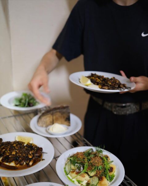 Osoba w czarnej koszulce trzyma talerz z roślinnym daniem, a na metalowej półce obok stoją inne talerze z wegańskimi potrawami, takimi jak gnocchi z ziołami, grzyby z sosem oraz chrupiący chleb z masłem roślinnym. Zdjęcie przedstawia kreatywne podejście do kuchni roślinnej w kontekście "najlepsze wegańskie restauracje warszawa 2025".