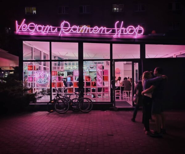 Fasada restauracji Vegan Ramen Shop oświetlona różowymi neonami, z dużymi oknami ukazującymi nowoczesne, jasne wnętrze pełne gości. Przed lokalem stoją rowery, a na chodniku obejmuje się para. Zdjęcie przedstawia popularne miejsce na kulinarnej mapie najlepsze wegańskie restauracje w Warszawie 2025.