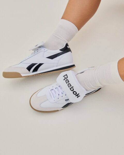 Zbliżenie na białe sneakersy Reebok z czarnymi detalami – Stylowe, białe buty sportowe Reebok z czarnymi paskami, gumową podeszwą i szerokim paskiem na rzep z logo marki, noszone z kremowymi skarpetkami.