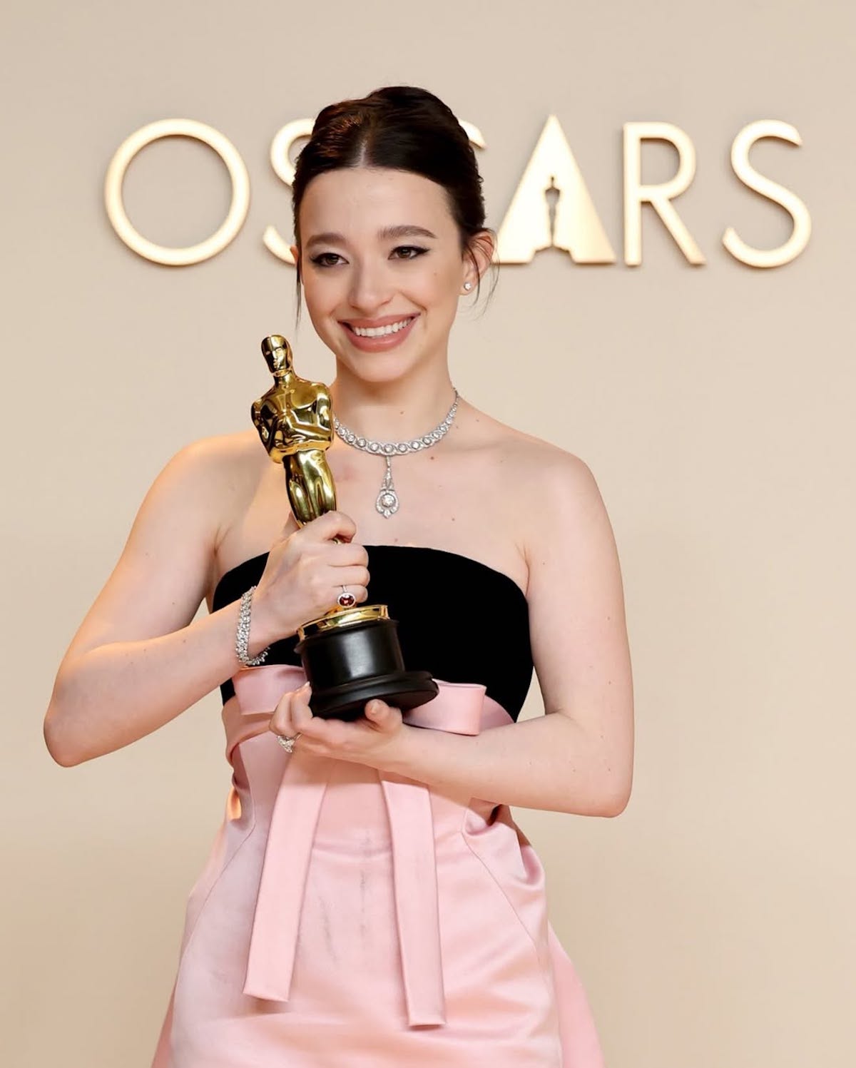 Zwyciężczyni Oscara 2025 pozuje z nagrodą na tle napisu „OSCARS”. Ubrana w elegancką suknię w kolorze różowym i czarnym, z diamentową biżuterią, trzyma statuetkę i uśmiecha się promiennie.