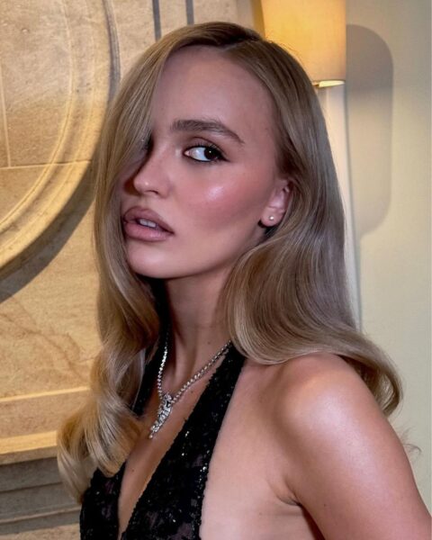 Zdjęcie aktorki Lily-Rose Depp. Ma rozpuszczone włosy na ramiona. Ubrana jest w czarną sukienkę na ramiączkach ozdobiona cekinami. Na szyi ma srebrny naszyjnik. Pozuje półbokiem na tle marmurowej ściany.