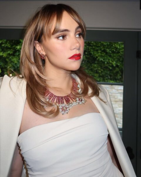 Zdjęcie Suki Waterhouse. Ma blond włosy rozjaśnione refleksami i grzywkę. Na szyi ma masywny naszyjnik z czerwonymi kamieniami i kwiatami. Ubrana jest w białą sukienkę bez ramiączkach, a na ramionach ma kremową marynarkę. Patrzy w bok. Znajduje się w pomieszczeniu, na tle okna i widać przez nie zielony żywopłot.