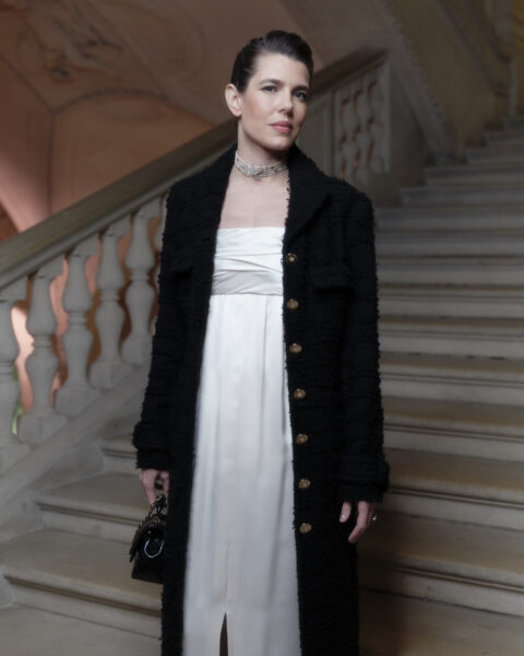 Charlotte Casiraghi pozuje na schodach podczas paryskiej Met Gali 2025. Kobieta ma na sobie długą białą sukienkę i czarny płaszcz o ciekawej fakturze. Na szyi ma srebrny choker, a w ręku trzyma małą czarną torebkę.