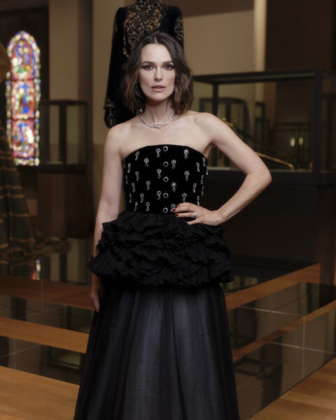 Keira Knightley pozuje podczas europejskiej Met Gali. Aktorka ma na sobie czarną suknię wieczorową, z górą z biżuteryjnymi detalami oraz falbanami w okolicach talii. W tle widać ekspozycję LOUVRE COUTURE. Art and Fashion: Statement Pieces.