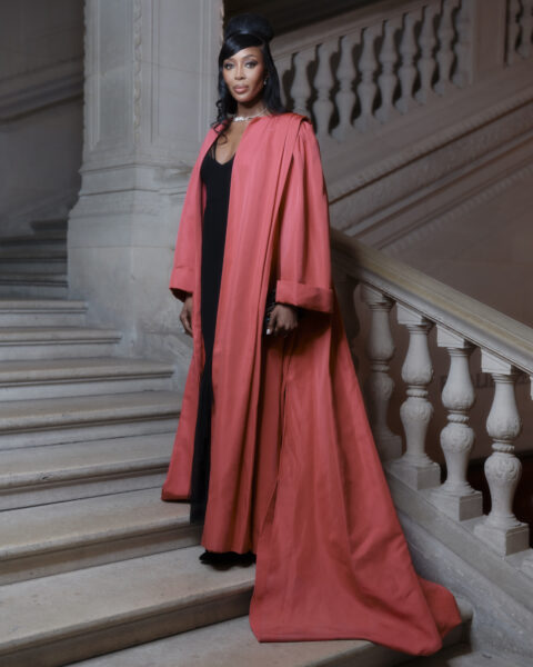Naomi Campbell w trakcie europejskiej Met Gali. Modelka ma na sobie długi, czerwony płaszcz i czarną sukienkę. Stoi na schodach i trzyma w ręku torebkę.