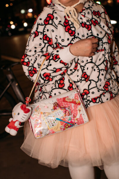 Stylizacja inspirowana kawaii; osoba w bluzie w motyw Hello Kitty, tiulowej spódnicy i białych rajstop trzyma przezroczystą torebkę wypełnioną japońskimi słodyczami i naklejkami, z breloczkiem w kształcie Hello Kitty.
