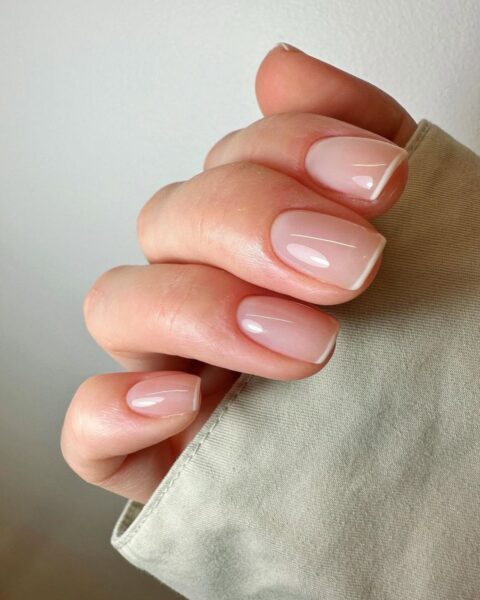Zdjęcie ze zbliżeniem na dłonie z krótkimi, kwadratowymi paznokciami. Zdobi je stylizacja w stylu micro french manicure, czyli z cienką, białą kreską na brzegu płytki.