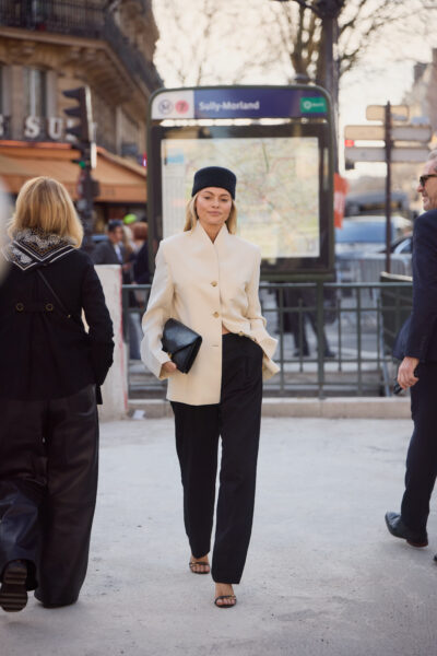 Trendy street style z Paryża: Minimalistyczna elegancja w miejskim wydaniu. Kobieta w kremowym oversizowym żakiecie z dużymi guzikami i czarnych szerokich spodniach. Stylizacja dopełniona czarną torebką kopertówką i elegancką czapką. Ulica Paryża w tle.