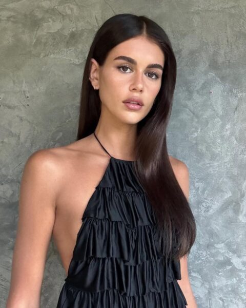 Modelka Kaia Gerber w długich, ciemnych włosach w kolorze brązowym. Są rozpuszczone i zaczesane na jedną stronę. Ubrana jest w czarny top z falbankami. Pozuje na tle betonowej ściany. 