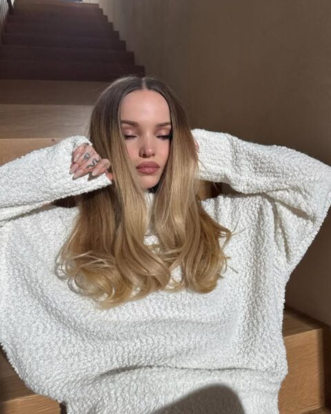 Modelka Dove Cameron w fryzurze typu ombre, która jest mocnym trendem w koloryzacji włosów. Ubrana jest w kremowoy sweter oversize oraz beżowe legginsy. Siedzi na stopniach schodów i przykłada ręce to warzy. 