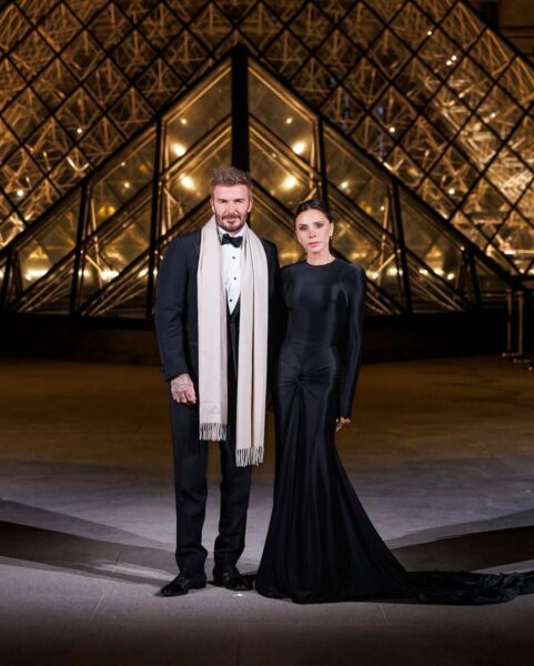 David i Victoria Beckham przed charytatywną kolacją z okazji europejskiej Met Gali 2025. Mężczyzna pozuje w smokingu i szaliku, a kobieta w długiej czarnej sukni z marszczeniem w okolicach brzucha. W tle widać rozświetlony Luwr.