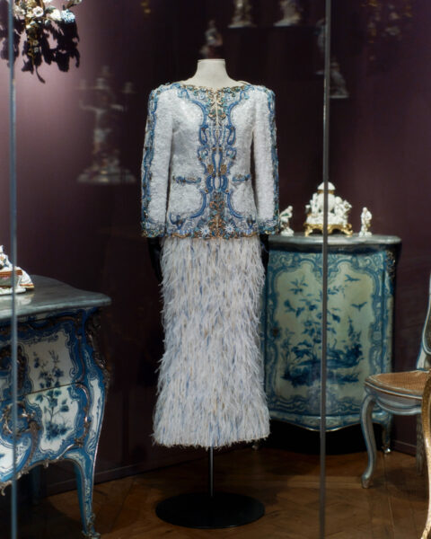 Elegancka marynarka CHANEL z misternym niebiesko-złotym haftem i piórkową spódnicą, prezentowana w stylizowanym, vintage’owym otoczeniu w trakcie LOUVRE COUTURE. Art and Fashion: Statement Pieces.