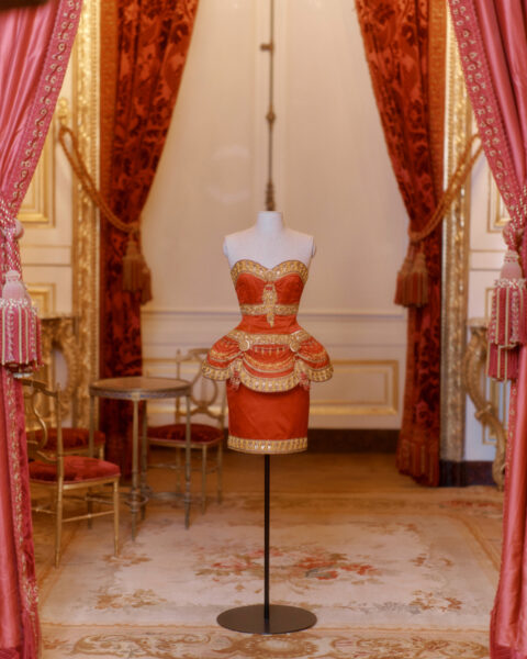 Piękna czerwona sukienka CHANEL ze złotymi zdobieniami, baskinką i gorsetową górą, wystawiona na manekinie w eleganckim pokoju, ozdobionym bogatymi zasłonami i vintage’owym meblami.