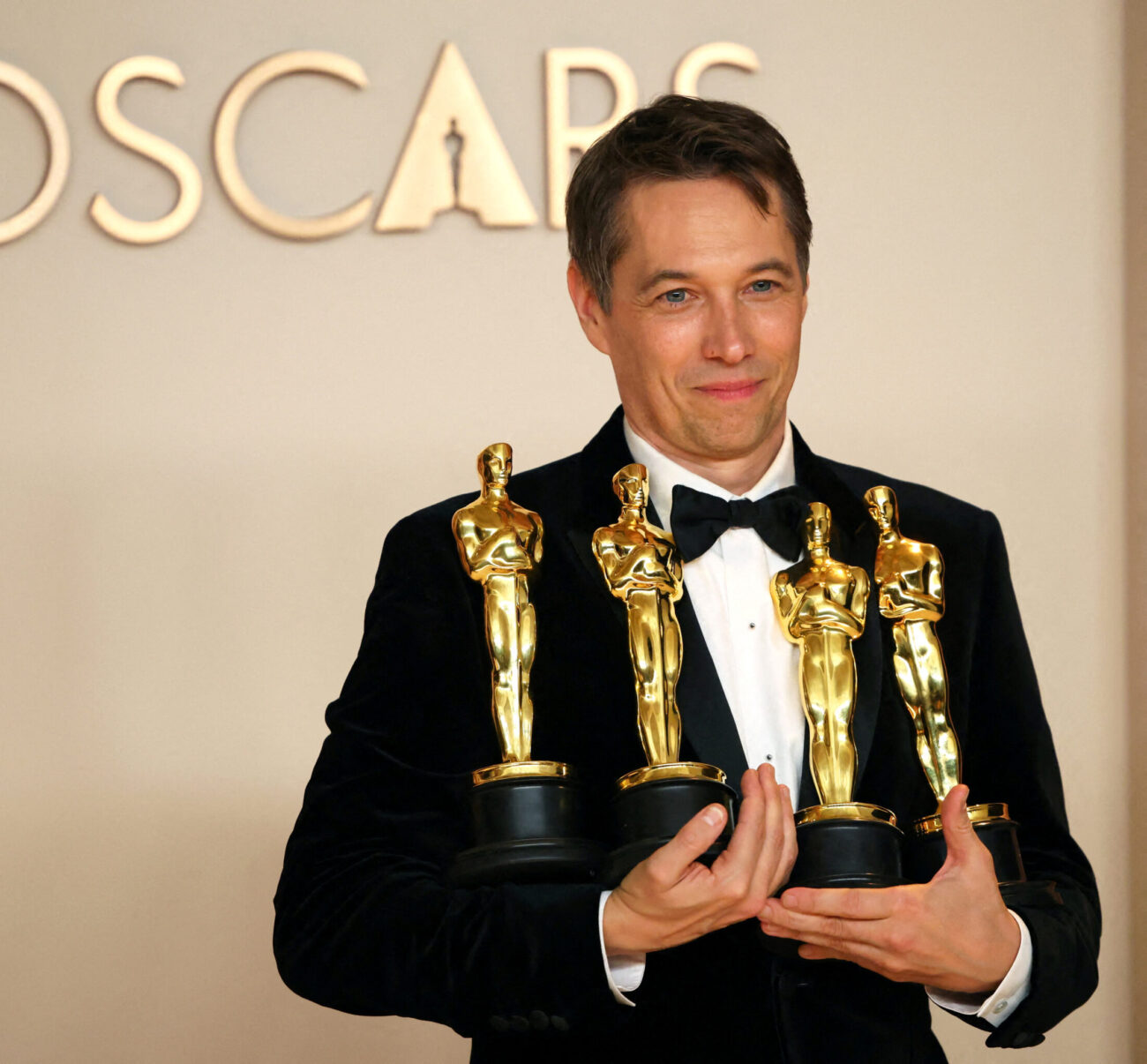 Mężczyzna w eleganckim czarnym smokingu i muszce stoi na tle napisu „OSCARS” i uśmiecha się, trzymając w ramionach cztery złote statuetki Oscara.