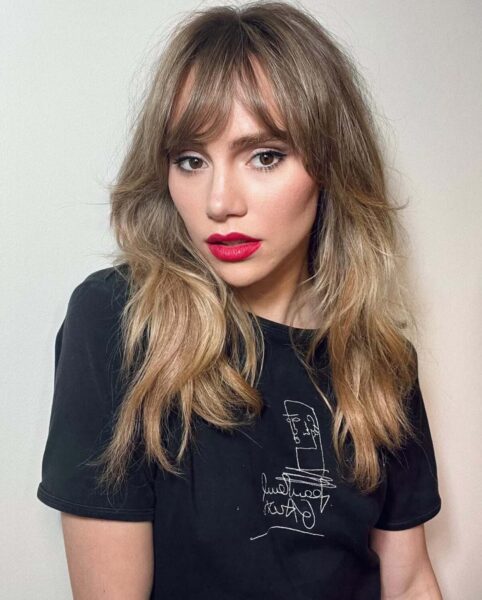 Delikatna grzywka na wiosnę 2025: Suki Waterhouse. Zdjęcie przedstawia kobietę w rozpuszczonych włosach i czarnej koszulce z nadrukiem. Kobieta ma usta podkreślone czerwoną szminką i fryzurę bottleneck bangs.