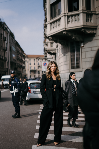 Trendy street style z Mediolanu: Total black look na ulicach miasta – Kobieta w czarnym garniturze z szerokimi spodniami i krótkim topem spaceruje po przejściu dla pieszych. Jej elegancka stylizacja jest uzupełniona przez czarne rękawiczki i minimalistyczną torebkę.