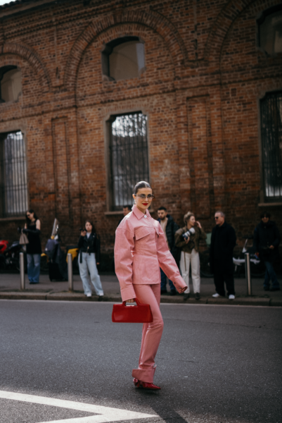 Trendy street style z Mediolanu: Różowy monochromatyczny look – Kobieta w pastelowo-różowym garniturze składającym się z oversize’owej kurtki i dopasowanych spodni. W ręku trzyma małą czerwoną torebkę, a jej look dopełniają okulary przeciwsłoneczne i czerwone usta.