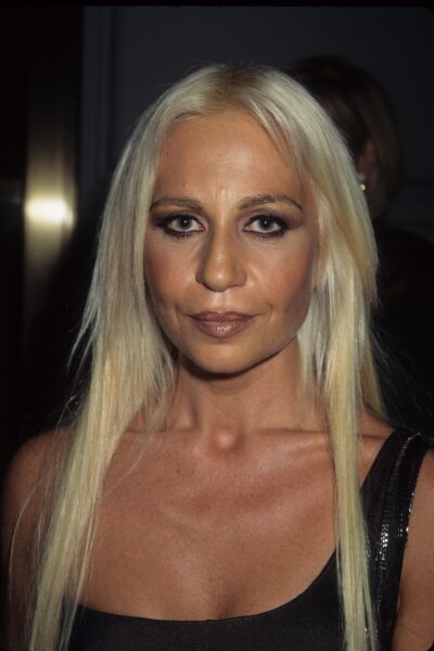 Młoda Donatella Versace. Kobieta ma lekki uśmiech na twarzy, blond włosy i mocno podkreślone oczy.