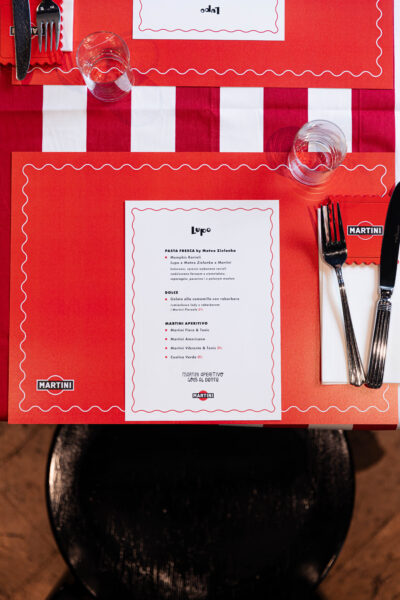 Nakrycie stołu w restauracji Lupo z kartą menu zaprojektowaną w stylistyce włoskiej trattorii – czerwono-białe obrusy, szkło i sztućce ułożone obok kartki z logotypami Lupo i Martini.
